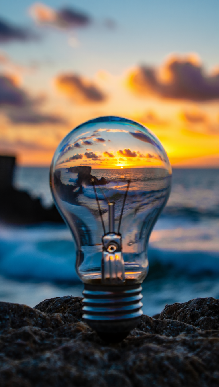 Coastal sunset through a clear glass light bulb positioned on coastal rocks