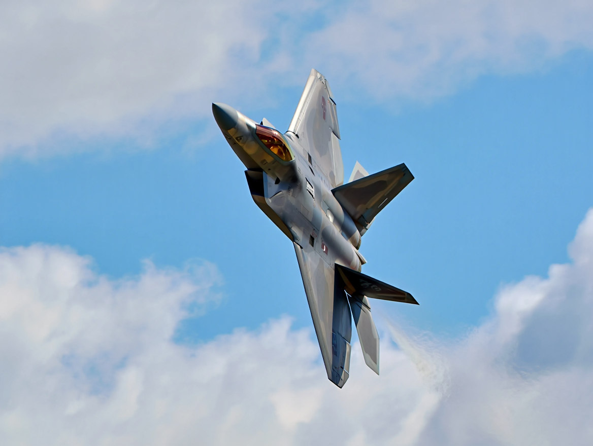 A F-22 Raptor military fighter jet with metallic grey fuselage soars through a bright blue sky filled with white cumulus clouds